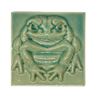 Könnte beinhalten: Eine quadratische Keramikfliese mit einem erhabenen Relief eines Frosches in einer glänzenden, hellen meergrünen Glasur. Der Frosch ist mittig auf der Fliese platziert, wobei die Vorderbeine nach vorne gestreckt sind und eine glatte Oberfläche zeigen.