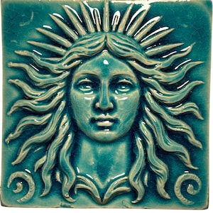 Op de afbeelding: Vierkante keramische tegel met een turquoise glazuur met een gedetailleerd reliëf van een zon-gezicht figuur. Het gezicht heeft serene kenmerken, omgeven door golvend haar en zonnestralen. Decoratieve krullen bevinden zich in de hoeken.