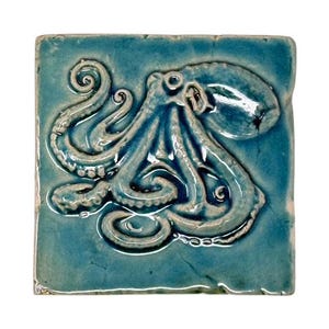 Könnte beinhalten: Quadratische Keramikfliese mit einem erhabenen Oktopus-Design. Der Oktopus ist in Hellblau gehalten, mit einer dunkleren blauen Glasur. Die Fliese hat eine glänzende Oberfläche und ein leichtes Used-Aussehen, was auf handwerkliche Qualität hindeutet. Die Fliese ist ein Dekorationsartikel.