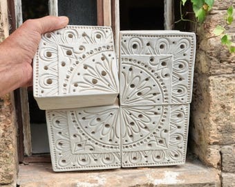 May include: Four square, off-white ceramic tiles with an intricate, carved floral and geometric design. The tiles are arranged together, with one held in a hand, showcasing the detailed pattern and texture. The tiles are displayed near a window.