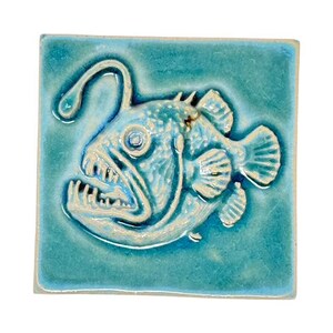 Könnte beinhalten: Quadratische, türkisfarbene Keramikfliese mit einem detaillierten Relief eines Anglerfischs. Der Fisch hat ein großes Maul mit scharfen Zähnen, einen leuchtenden Köder und strukturierte Flossen. Dekorativer Artikel.