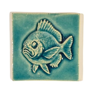 Könnte beinhalten: Eine quadratische, türkisfarbene Keramikfliese mit einem detaillierten Relief eines Fisches. Der Fisch hat strukturierte Schuppen und Flossen, mit einem markanten Auge und Mund. Die Fliese hat eine glänzende Oberfläche und einen weißen Rand.