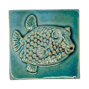 Könnte beinhalten: Quadratische Keramikfliese mit türkisfarbener Glasur, die ein erhabenes Design eines Kugelfisches zeigt. Der Fisch hat einen strukturierten Körper mit kleinen, runden Unebenheiten und detaillierten Flossen. Die Fliese ist ein dekoratives Element.