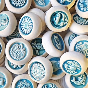 Könnte beinhalten: Eine Sammlung weißer Keramiksteine mit verschiedenen blauen Designs. Die Designs umfassen ein Seepferdchen, eine Blume, einen Fisch, einen Fuchs und andere Meereskreaturen. Die Steine sind rund und haben ein erhabenes, strukturiertes Aussehen.