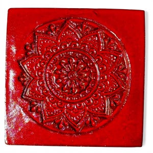 Könnte beinhalten: Quadratische, rote Keramikfliese mit einem erhabenen, kreisförmigen Blumendesign in der Mitte. Die Fliese hat eine glänzende Oberfläche und eine leicht strukturierte Oberfläche. Das Design zeigt ein symmetrisches Muster aus Blütenblättern und anderen dekorativen Elementen.