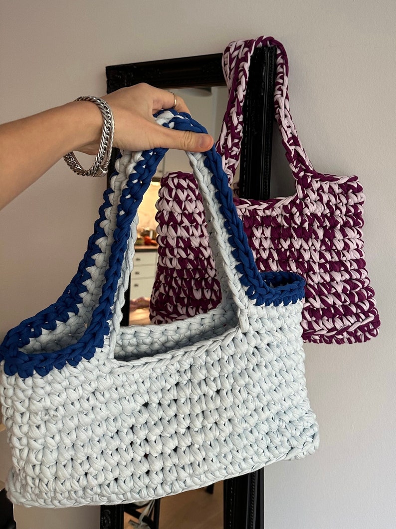 May include: Two crocheted tote bags, one light blue with a navy blue trim and one purple with a pink trim.