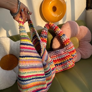 K&ouml;nnte beinhalten: Zwei handgefertigte H&auml;keltaschen mit bunten Streifenmustern. Die Taschen weisen eine Vielzahl leuchtender Farben auf, darunter Rosa, Blau, Gelb und Gr&uuml;n. Die Taschen werden von einer Person gehalten. Der Hintergrund umfasst ein kissenf&ouml;rmiges Kissen und eine donut-f&ouml;rmige Wanddekoration.