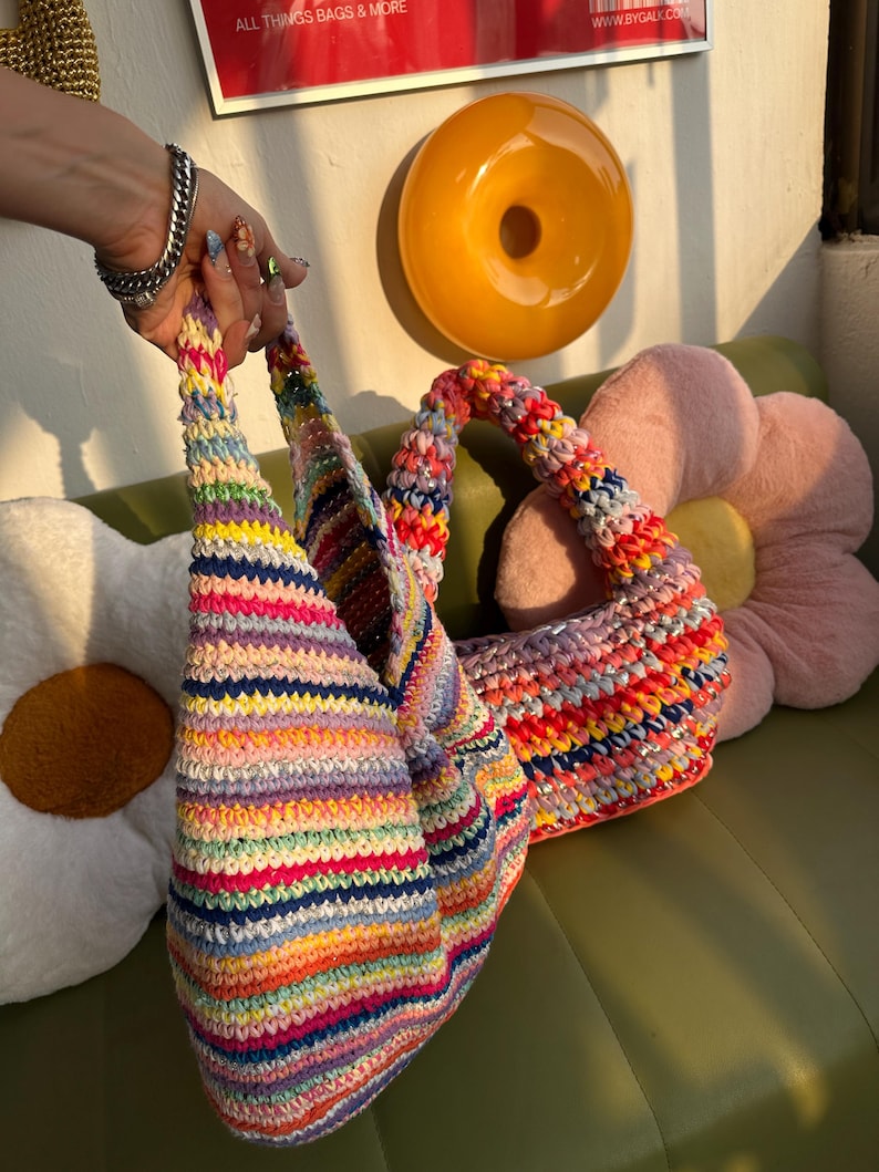 K&ouml;nnte beinhalten: Zwei handgefertigte H&auml;keltaschen mit bunten Streifenmustern. Die Taschen weisen eine Vielzahl leuchtender Farben auf, darunter Rosa, Blau, Gelb und Gr&uuml;n. Die Taschen werden von einer Person gehalten. Der Hintergrund umfasst ein kissenf&ouml;rmiges Kissen und eine donut-f&ouml;rmige Wanddekoration.