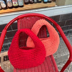 May include: Two crocheted bags, one red and one coral, are sitting on a red wicker chair. The bags are made with a simple stitch and have a rounded shape.