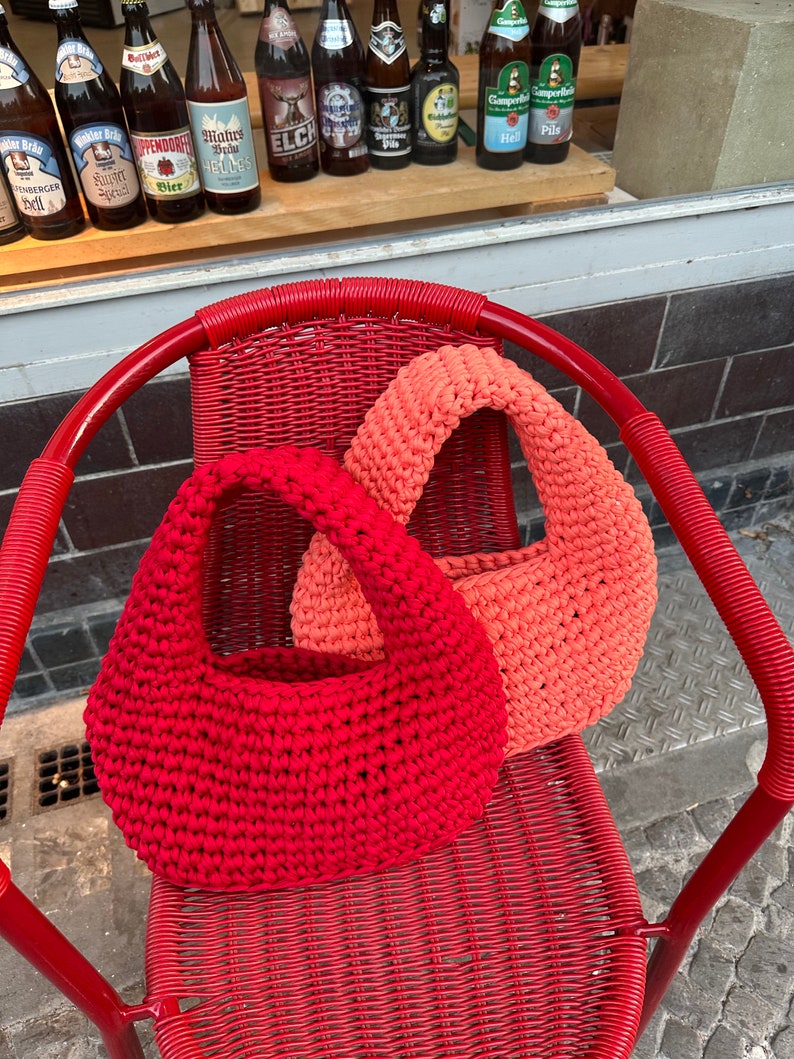 May include: Two crocheted bags, one red and one coral, are sitting on a red wicker chair. The bags are made with a simple stitch and have a rounded shape.