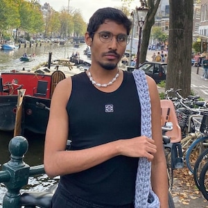 K&ouml;nnte beinhalten: Eine Person tr&auml;gt ein schwarzes Tanktop mit einem wei&szlig;en Logo, eine Perlenkette und eine hellblaue Stricktasche. Die Person steht vor einem Kanal mit Booten und B&auml;umen im Hintergrund.