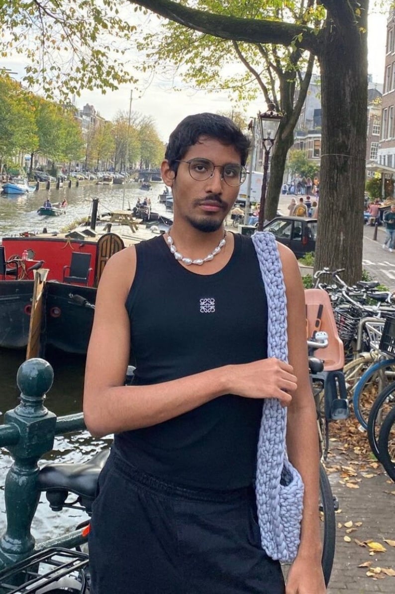 K&ouml;nnte beinhalten: Eine Person tr&auml;gt ein schwarzes Tanktop mit einem wei&szlig;en Logo, eine Perlenkette und eine hellblaue Stricktasche. Die Person steht vor einem Kanal mit Booten und B&auml;umen im Hintergrund.