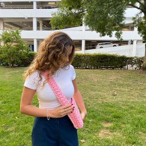 K&ouml;nnte beinhalten: Eine rosa geh&auml;kelte Schultertasche, die &uuml;ber einem wei&szlig;en T-Shirt und einer dunkelblauen Hose getragen wird.