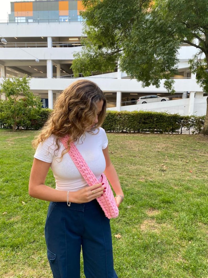 K&ouml;nnte beinhalten: Eine rosa geh&auml;kelte Schultertasche, die &uuml;ber einem wei&szlig;en T-Shirt und einer dunkelblauen Hose getragen wird.