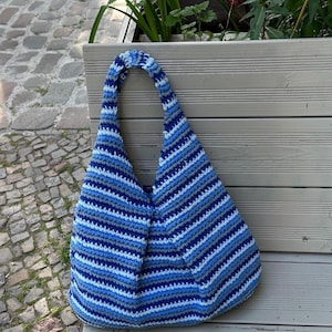 K&ouml;nnte beinhalten: Eine blau-wei&szlig; gestreifte H&auml;keltasche mit breitem Tr&auml;ger. Die Tasche ist aus weichem Garn gefertigt und hat einen abgerundeten Boden. Die Tasche liegt auf einer Holzbank.