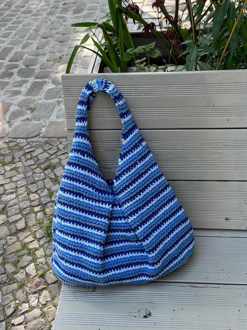 K&ouml;nnte beinhalten: Eine blau-wei&szlig; gestreifte H&auml;keltasche mit breitem Tr&auml;ger. Die Tasche ist aus weichem Garn gefertigt und hat einen abgerundeten Boden. Die Tasche liegt auf einer Holzbank.