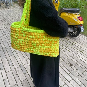 May include: A large, crocheted tote bag with a bright yellow and green color scheme. The bag has a wide, flat base and two thick, crocheted straps.