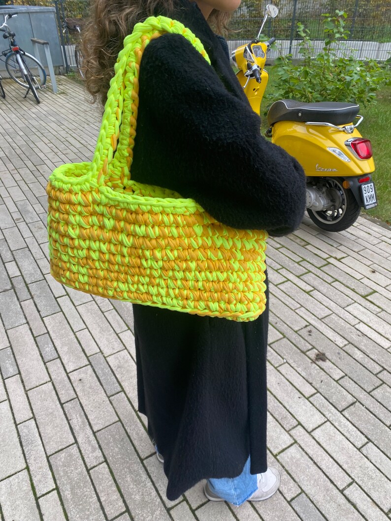 May include: A large, crocheted tote bag with a bright yellow and green color scheme. The bag has a wide, flat base and two thick, crocheted straps.