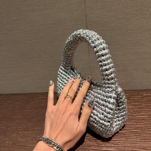 Pu&ograve; includere: Una piccola borsa a mano all'uncinetto color argento con un manico arrotondato. La borsa ha un aspetto strutturato e un design compatto, adatto per trasportare l'essenziale. La borsa &egrave; realizzata con un filato lucido e metallico.