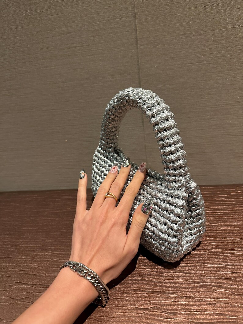 Pu&ograve; includere: Una piccola borsa a mano all'uncinetto color argento con un manico arrotondato. La borsa ha un aspetto strutturato e un design compatto, adatto per trasportare l'essenziale. La borsa &egrave; realizzata con un filato lucido e metallico.