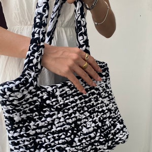 May include: A black and white crocheted tote bag with thick straps. The bag is made from recycled materials and has a unique, textured design.