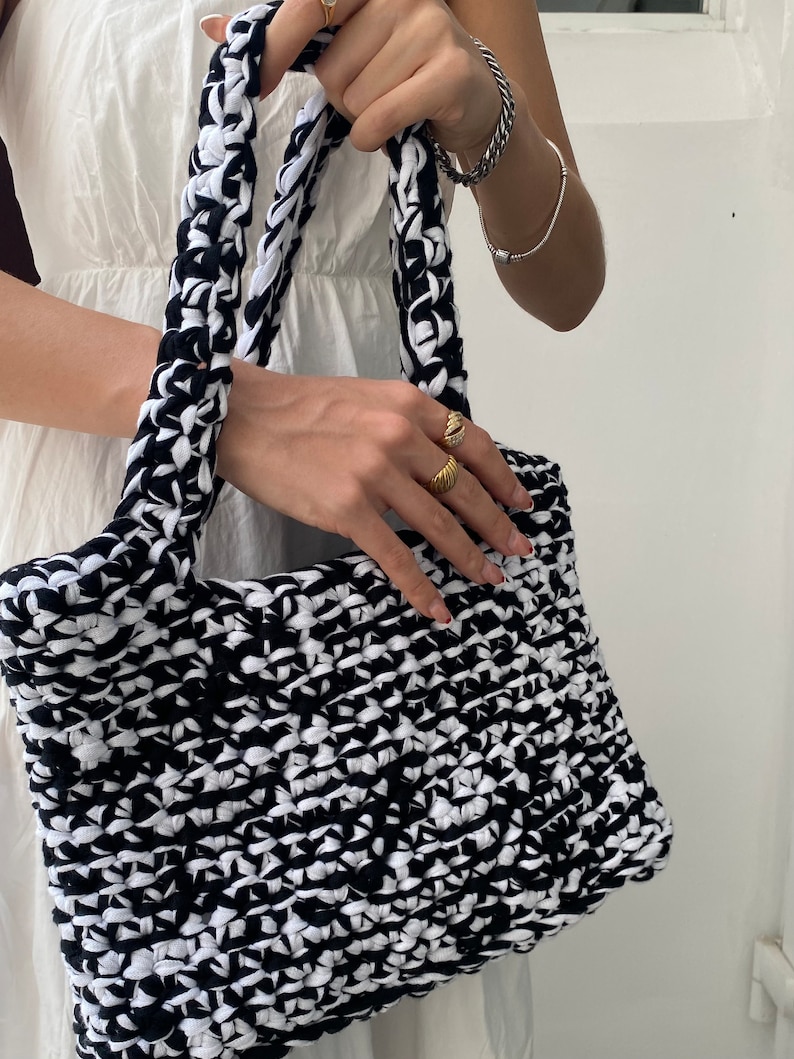 May include: A black and white crocheted tote bag with thick straps. The bag is made from recycled materials and has a unique, textured design.