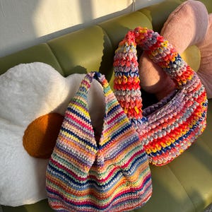 Puede incluir: Dos bolsos de crochet hechos a mano en vibrantes colores arco&iacute;ris. Uno es un bolso tote con una abertura ancha y un patr&oacute;n de rayas. El otro es un bolso de hombro con forma redondeada y una correa gruesa. Ambos bolsos est&aacute;n sobre una superficie verde.