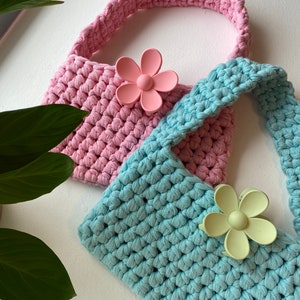 May include: Two crocheted bags with flower-shaped clips. The bag on top is pink and the bag on the bottom is light blue.