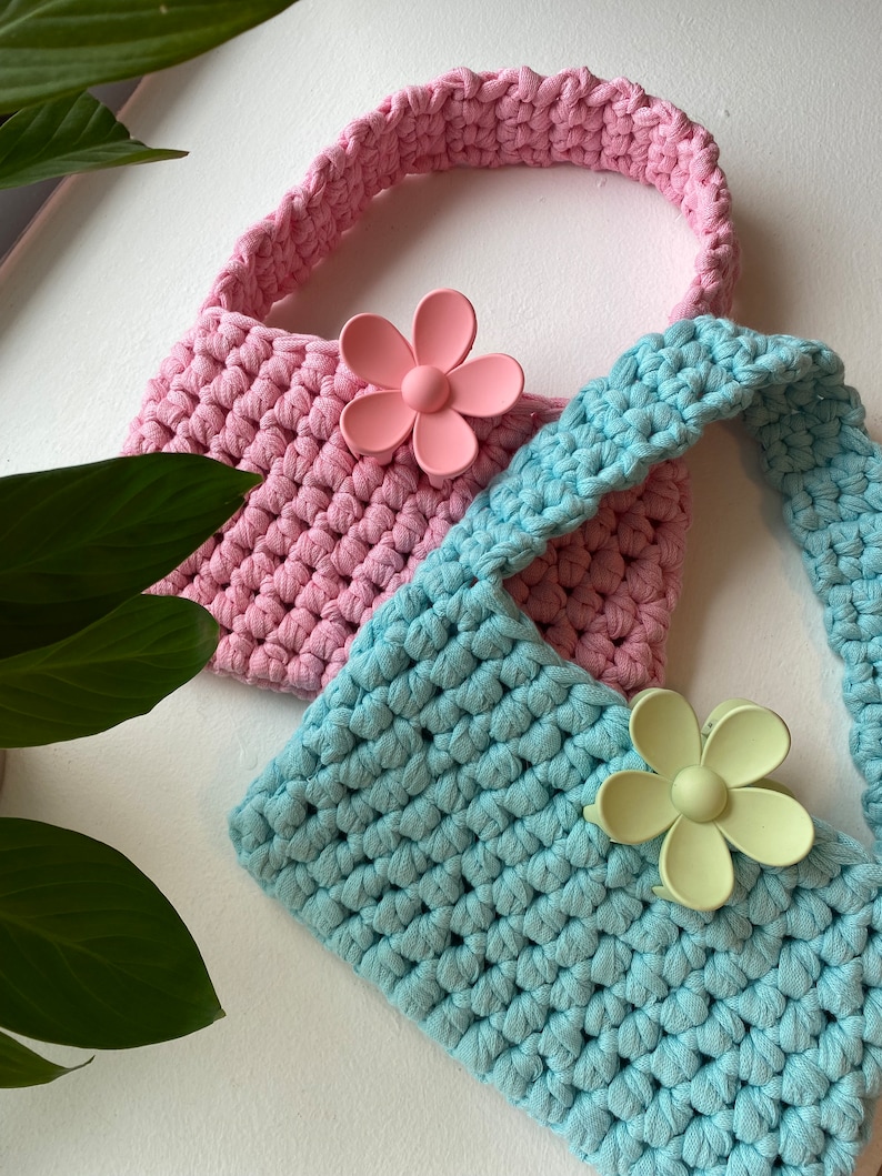 May include: Two crocheted bags with flower-shaped clips. The bag on top is pink and the bag on the bottom is light blue.