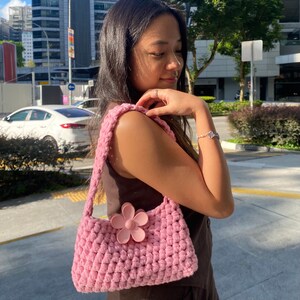 May include: A pink crocheted handbag with a large pink flower accent. The bag has a long strap and is worn over the shoulder.