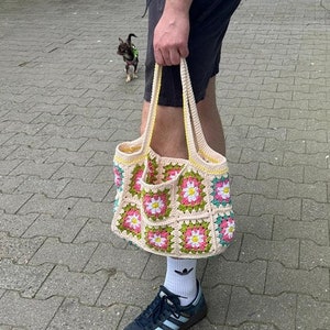 Op de afbeelding: Een gehaakte tote bag met een bloemenpatroon in roze, wit en groen. De tas heeft een gele bies en is gemaakt van een zacht, getextureerd garen.