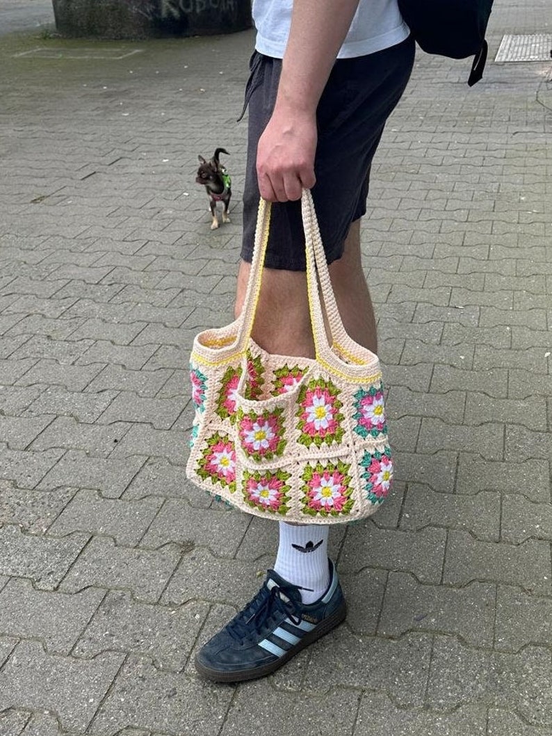 Op de afbeelding: Een gehaakte tote bag met een bloemenpatroon in roze, wit en groen. De tas heeft een gele bies en is gemaakt van een zacht, getextureerd garen.