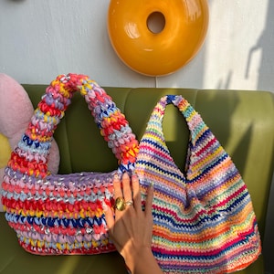 Puede incluir: Dos coloridos bolsos de crochet hechos a mano. Uno tiene forma de media luna con un arco&iacute;ris de colores y lentejuelas plateadas. El otro tiene forma triangular con rayas horizontales de varios colores. Ambos bolsos tienen asas gruesas y texturizadas.