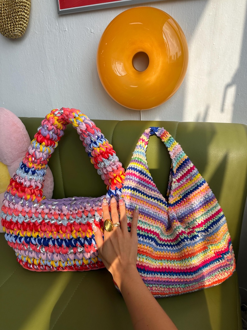 Puede incluir: Dos coloridos bolsos de crochet hechos a mano. Uno tiene forma de media luna con un arco&iacute;ris de colores y lentejuelas plateadas. El otro tiene forma triangular con rayas horizontales de varios colores. Ambos bolsos tienen asas gruesas y texturizadas.