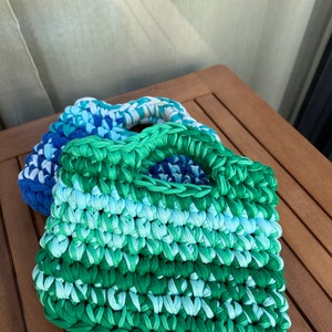 May include: A crocheted tote bag with a green, blue, and white color gradient. The bag has a single handle and is made from a soft, textured yarn.