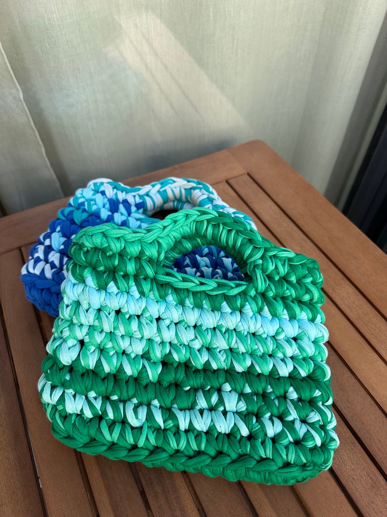 May include: A crocheted tote bag with a green, blue, and white color gradient. The bag has a single handle and is made from a soft, textured yarn.