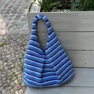 Puede incluir: Bolso tote de crochet hecho a mano con rayas azules y blancas. El bolso tiene forma redondeada y un asa &uacute;nica. Descansa sobre un banco de madera, perfecto para llevar lo esencial.
