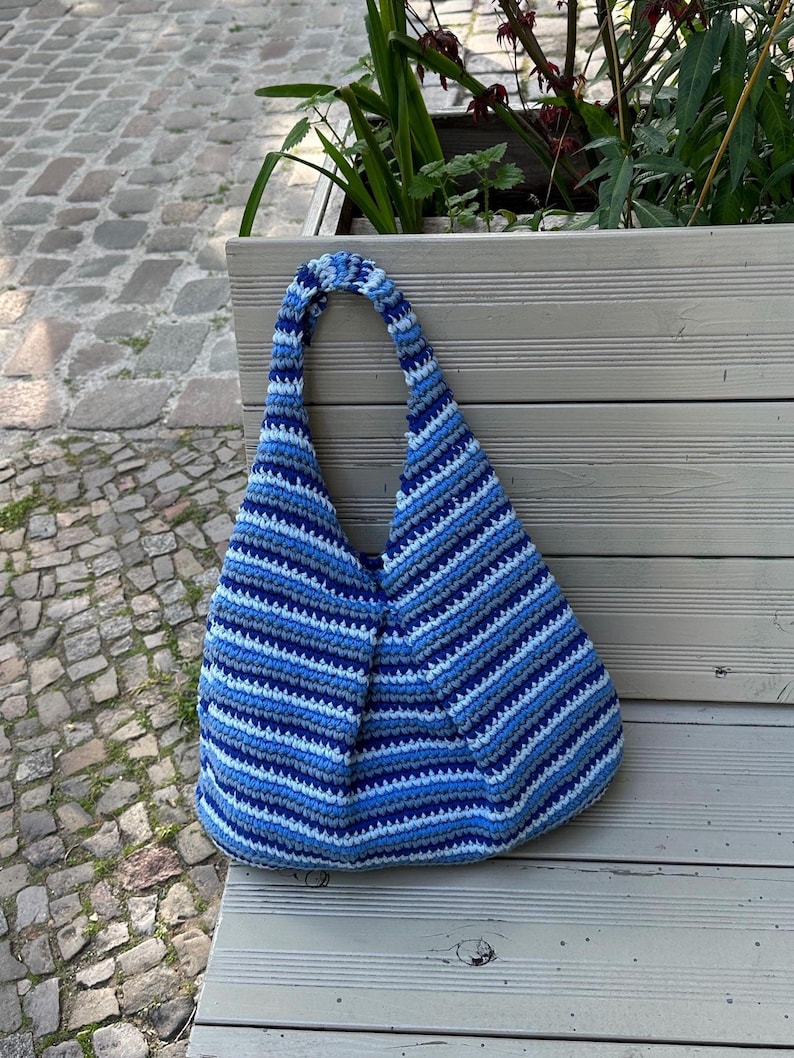 Puede incluir: Bolso tote de crochet hecho a mano con rayas azules y blancas. El bolso tiene forma redondeada y un asa &uacute;nica. Descansa sobre un banco de madera, perfecto para llevar lo esencial.