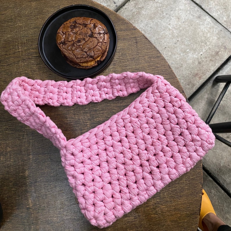 May include: A pink crocheted bag with a thick strap. The bag is made of a soft, textured yarn and has a simple, classic design.