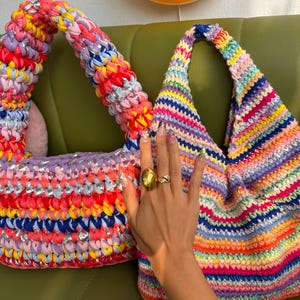 K&ouml;nnte beinhalten: Zwei farbenfrohe, handgefertigte H&auml;keltaschen. Eine ist eine Tragetasche mit breitem Griff, in Pink, Rot, Gelb und Blau. Die andere ist eine dreieckige Tasche mit Regenbogenmuster. Beide Taschen liegen auf einer gr&uuml;nen Oberfl&auml;che.