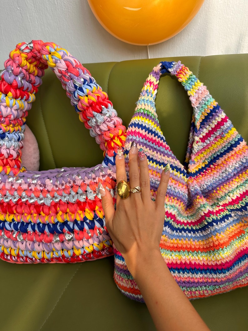 K&ouml;nnte beinhalten: Zwei farbenfrohe, handgefertigte H&auml;keltaschen. Eine ist eine Tragetasche mit breitem Griff, in Pink, Rot, Gelb und Blau. Die andere ist eine dreieckige Tasche mit Regenbogenmuster. Beide Taschen liegen auf einer gr&uuml;nen Oberfl&auml;che.