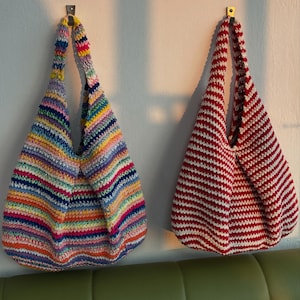 Puede incluir: Dos bolsos de mano de ganchillo hechos a mano colgados de una pared blanca. Un bolso presenta rayas horizontales de varios colores, mientras que el otro tiene rayas rojas y blancas. Ambos bolsos tienen forma redondeada y asas largas.