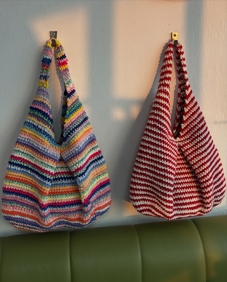 Puede incluir: Dos bolsos de mano de ganchillo hechos a mano colgados de una pared blanca. Un bolso presenta rayas horizontales de varios colores, mientras que el otro tiene rayas rojas y blancas. Ambos bolsos tienen forma redondeada y asas largas.
