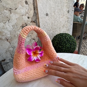Pu&ograve; includere: Una piccola borsa a mano all'uncinetto in tonalit&agrave; rosa e arancio con un accento di fiore rosa. La borsa ha un'unica maniglia ed &egrave; realizzata con un punto testurizzato.