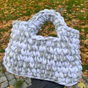 May include: A white and gray crocheted tote bag with a wide handle. The bag is made from thick yarn and has a textured, woven appearance.