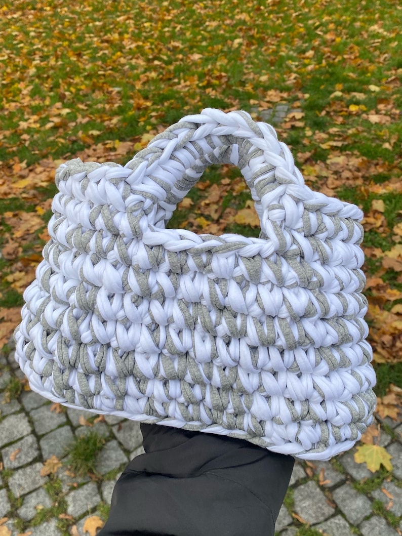 May include: A white and gray crocheted tote bag with a wide handle. The bag is made from thick yarn and has a textured, woven appearance.