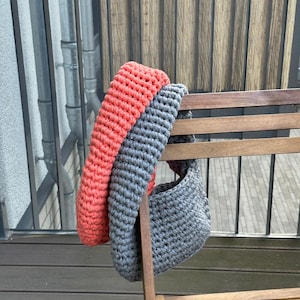 May include: A gray and coral crocheted tote bag hanging on a wooden chair. The bag is made of thick yarn and has a simple, rounded shape.