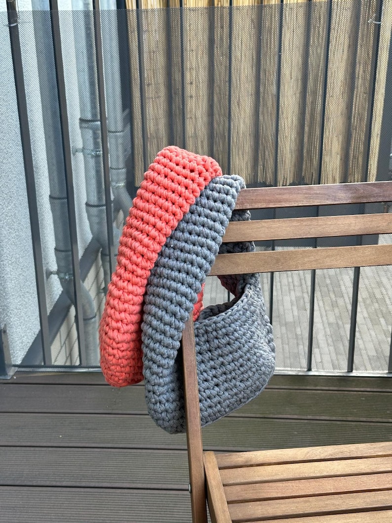 May include: A gray and coral crocheted tote bag hanging on a wooden chair. The bag is made of thick yarn and has a simple, rounded shape.
