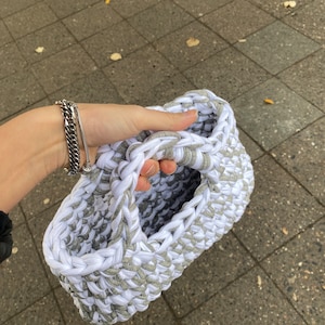 May include: A white and gray crocheted tote bag with a single handle. The bag is made from thick yarn and has a textured surface.