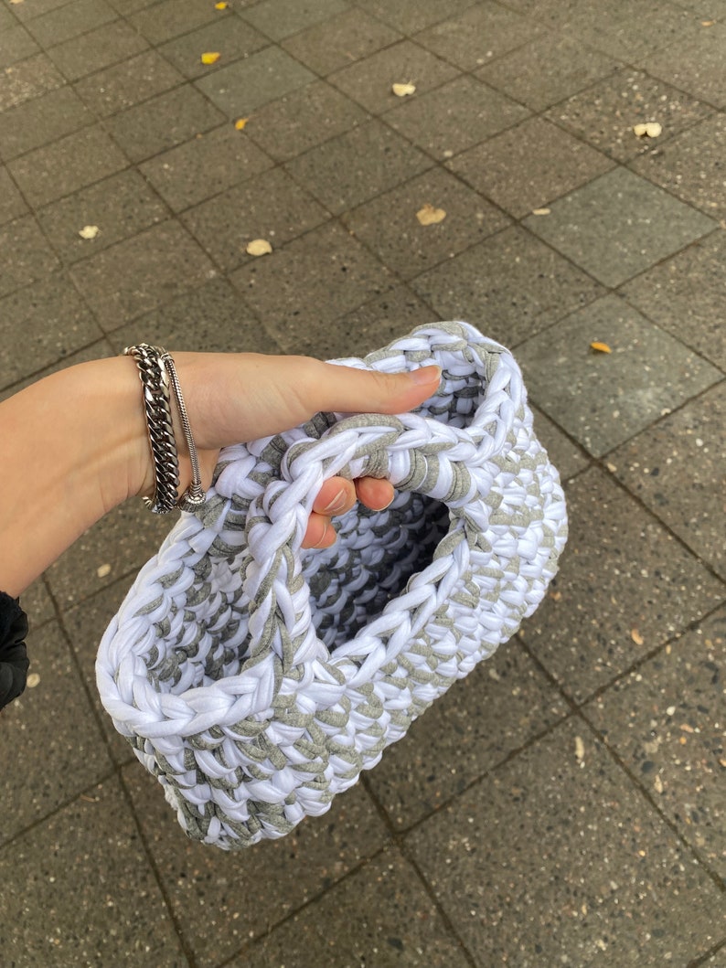 May include: A white and gray crocheted tote bag with a single handle. The bag is made from thick yarn and has a textured surface.